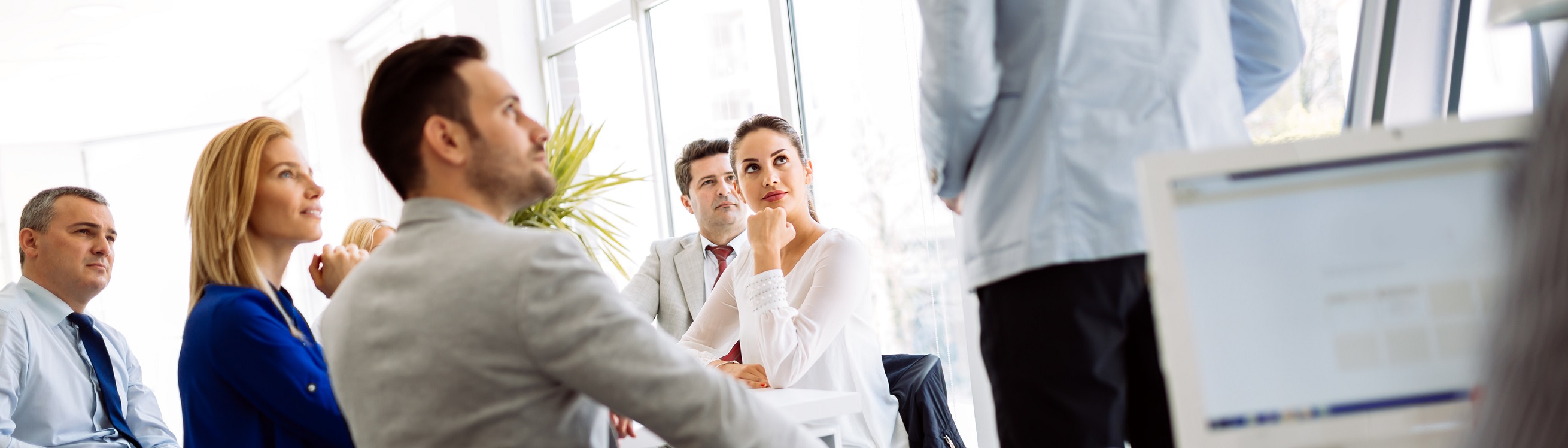 Business meeting with several white collar workers around a table, looking towards a presentation